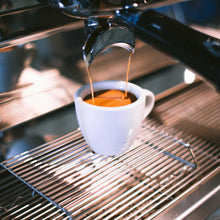 Load image into Gallery viewer, Milano Espresso Premium Blend die gezet wordt met een espressomachine. De koffie loopt uit de filterdrager in een espressokopje, waarin een mooie crema te zien is.
